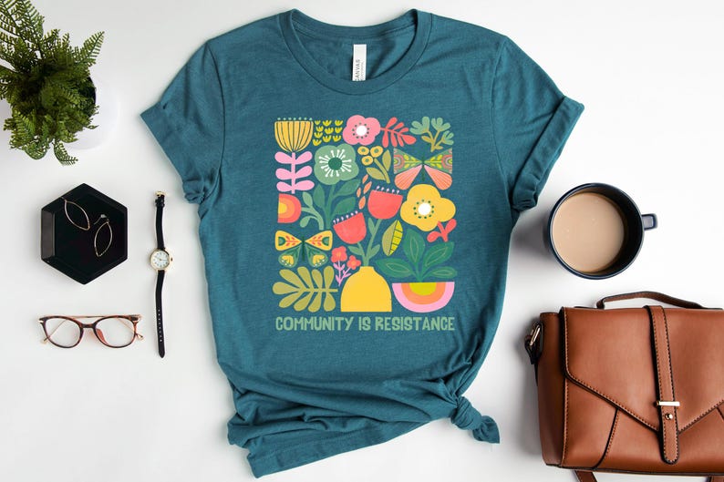 Op de afbeelding: Tealblauwe T-shirt met een kleurrijk bloemenontwerp en de tekst "Community is Resistance".