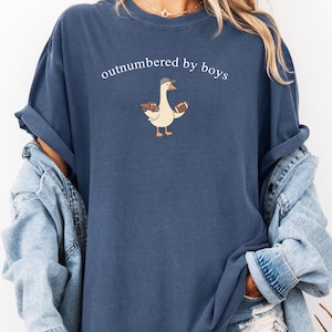 May include: A navy blue t-shirt with the text "outnumbered by boys". The shirt features a cartoon goose wearing a baseball cap and holding a football and a toy truck. The shirt is a relaxed fit.