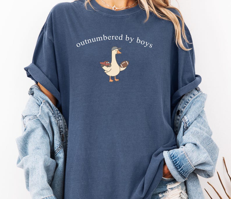 May include: A navy blue t-shirt with the text "outnumbered by boys". The shirt features a cartoon goose wearing a baseball cap and holding a football and a toy truck. The shirt is a relaxed fit.