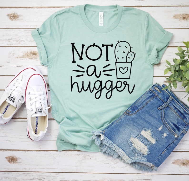 May include: A light green t-shirt with the text "NOT a hugger" and a black line drawing of a cactus in a pot. The shirt is laying on a white wooden surface with a pair of white canvas shoes and a pair of blue denim shorts.