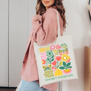 Op de afbeelding: Een witte tote bag met een kleurrijk bloemenontwerp en de tekst "Community is Resistance". De bloemen zijn roze, geel, rood, groen en blauw. De tas heeft een lange witte band.