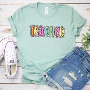 Puede incluir: Una camiseta azul claro con la palabra "teacher" en letras de colores con un patr&oacute;n de lunares blancos.