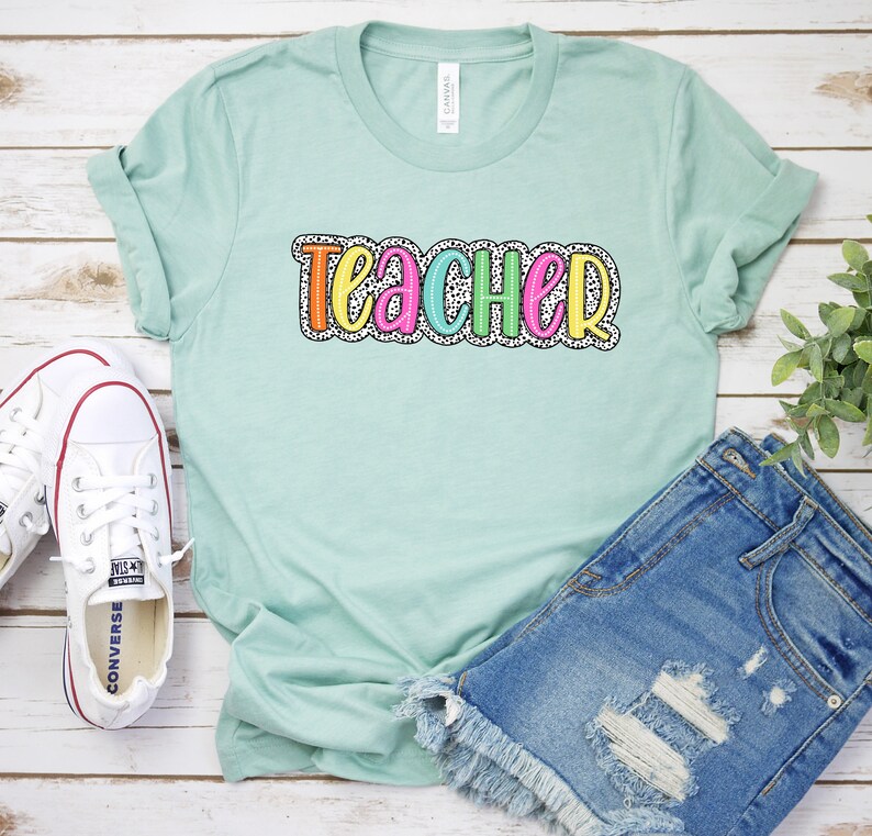 Puede incluir: Una camiseta azul claro con la palabra "teacher" en letras de colores con un patr&oacute;n de lunares blancos.