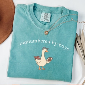 May include: Teal Comfort Colors t-shirt with the phrase "outnumbered by boys" in white. The shirt features a cartoon goose wearing a baseball cap and holding a football and a red truck. A gold necklace is draped across the shirt.