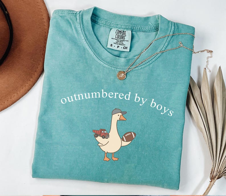May include: Teal Comfort Colors t-shirt with the phrase "outnumbered by boys" in white. The shirt features a cartoon goose wearing a baseball cap and holding a football and a red truck. A gold necklace is draped across the shirt.