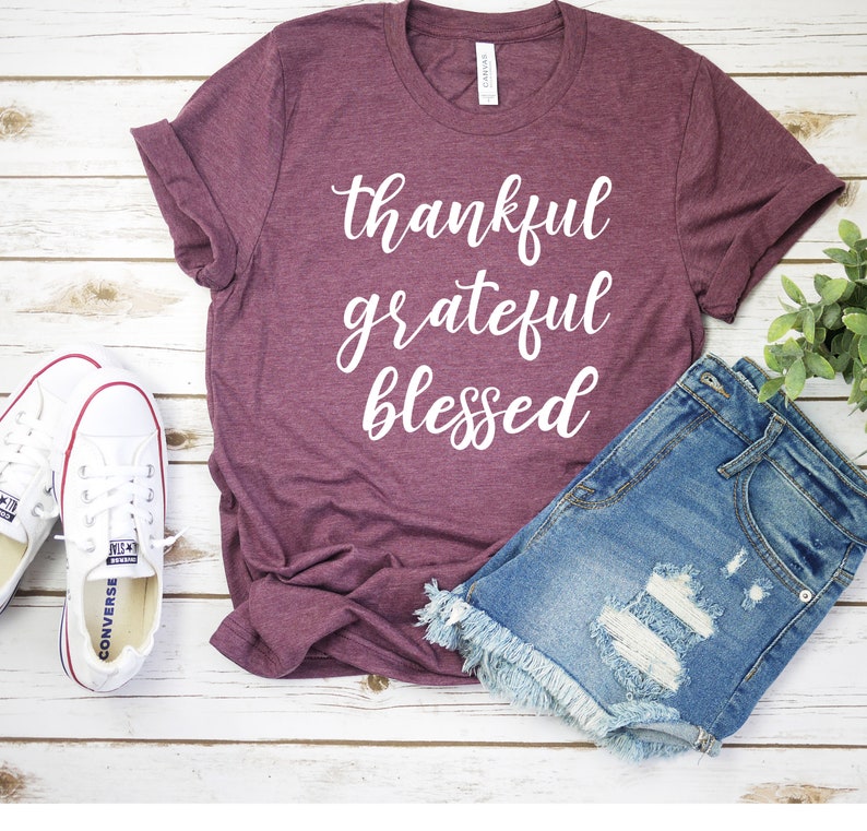 May include: A maroon t-shirt with the words "thankful grateful blessed" printed in white on the front. The shirt is laying on a white wooden surface with a pair of white sneakers and a pair of blue denim shorts.