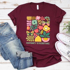 Op de afbeelding: Een maroon t-shirt met een kleurrijk bloemenontwerp en de tekst "Community is Resistance".