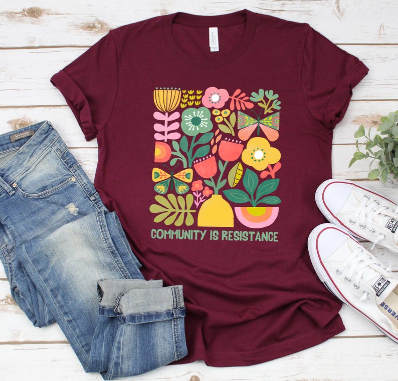 Op de afbeelding: Een maroon t-shirt met een kleurrijk bloemenontwerp en de tekst "Community is Resistance".