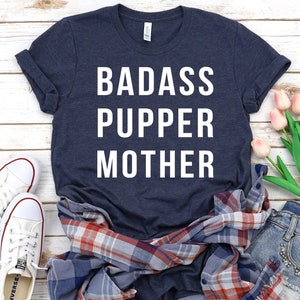 May include: A navy blue t-shirt with white text that reads "Badass Pupper Mother". The shirt is laid out on a white wooden surface with a plaid shirt, a pair of white sneakers, and a bouquet of pink tulips.