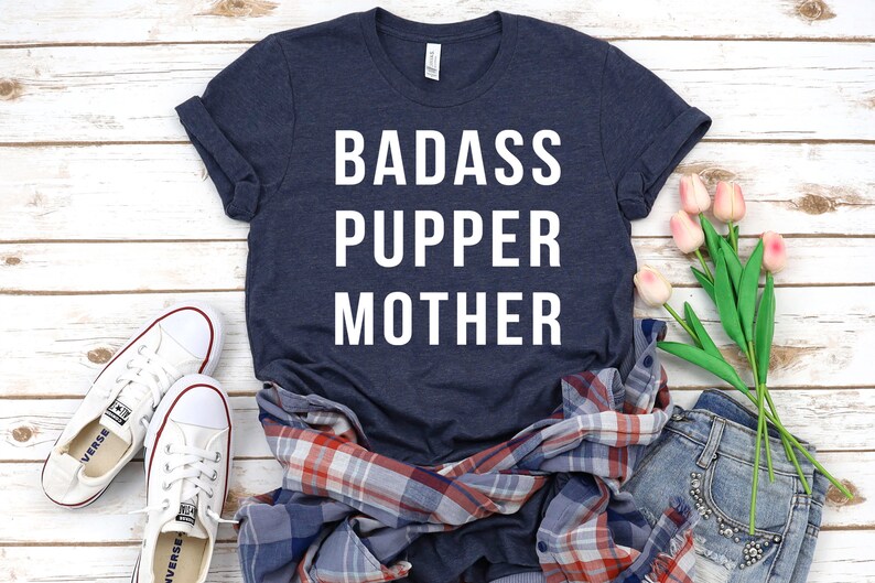 May include: A navy blue t-shirt with white text that reads "Badass Pupper Mother". The shirt is laid out on a white wooden surface with a plaid shirt, a pair of white sneakers, and a bouquet of pink tulips.