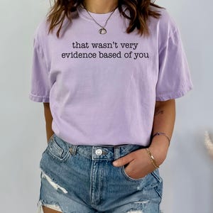 May include: A light purple t-shirt with the phrase "that wasn't very evidence based of you" printed in black. The shirt is paired with denim shorts.
