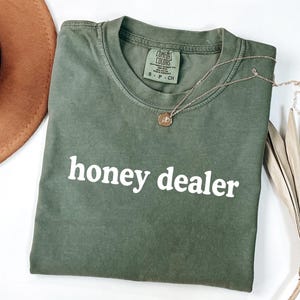 May include: An olive green t-shirt with the text "honey dealer" in white block letters. A gold-colored necklace with a small pendant rests on the shirt. A brown hat and dried plant are in the background.
