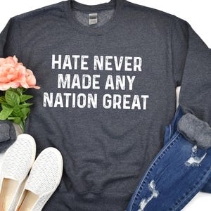 May include: A dark gray sweatshirt with the text "Hate Never Made Any Nation Great" printed in white on the front. The sweatshirt is laid flat on a white surface with a pair of white shoes and blue jeans.