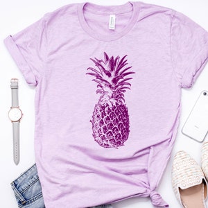Pode incluir: Uma camiseta de cor lil&aacute;s claro com um gr&aacute;fico de abacaxi roxo. A camiseta est&aacute; colocada sobre uma superf&iacute;cie branca com um rel&oacute;gio, um telefone e um par de sapatos brancos.