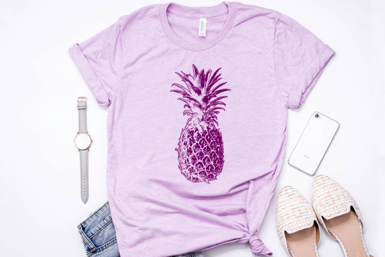 Pode incluir: Uma camiseta de cor lil&aacute;s claro com um gr&aacute;fico de abacaxi roxo. A camiseta est&aacute; colocada sobre uma superf&iacute;cie branca com um rel&oacute;gio, um telefone e um par de sapatos brancos.