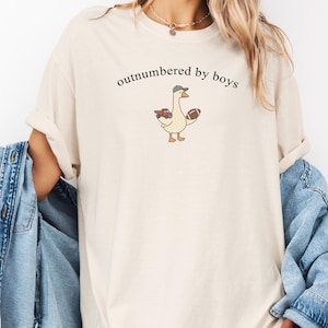 May include: Cream-coloured t-shirt featuring the text "outnumbered by boys" above a cartoon goose wearing a baseball cap and holding a football and a red toy truck. The shirt has rolled-up sleeves.