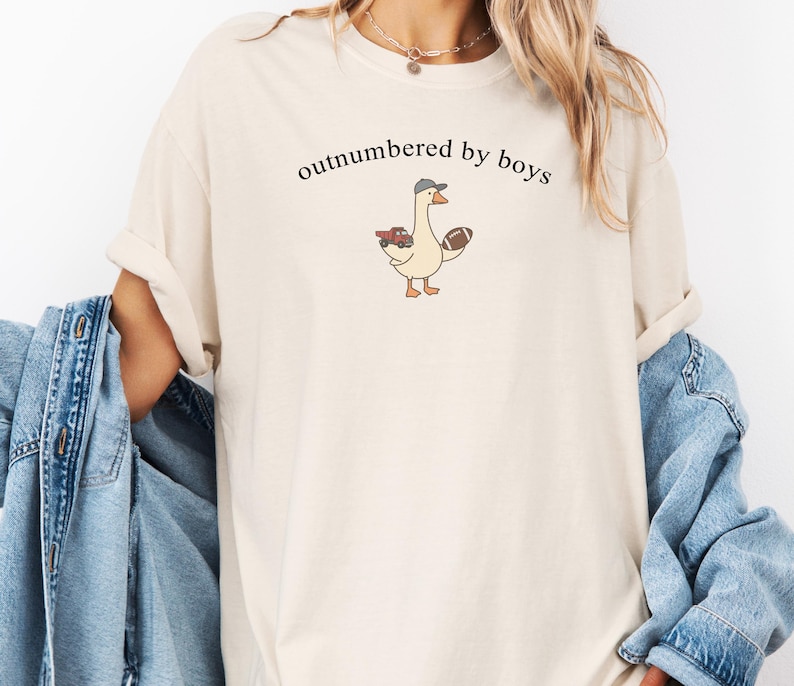 May include: Cream-coloured t-shirt featuring the text "outnumbered by boys" above a cartoon goose wearing a baseball cap and holding a football and a red toy truck. The shirt has rolled-up sleeves.