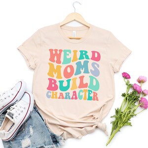 Pode incluir: Uma camiseta bege com o texto "Weird Moms Build Character" em cores do arco-&iacute;ris. O texto est&aacute; em um estilo de fonte retr&ocirc;.