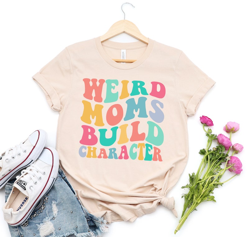 Pode incluir: Uma camiseta bege com o texto "Weird Moms Build Character" em cores do arco-&iacute;ris. O texto est&aacute; em um estilo de fonte retr&ocirc;.