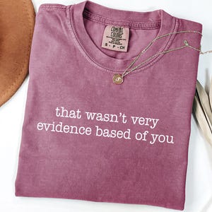 May include: A mauve Comfort Colors t-shirt with the phrase "that wasn't very evidence based of you" in white text. A gold necklace with a pendant and a brown hat are also visible.