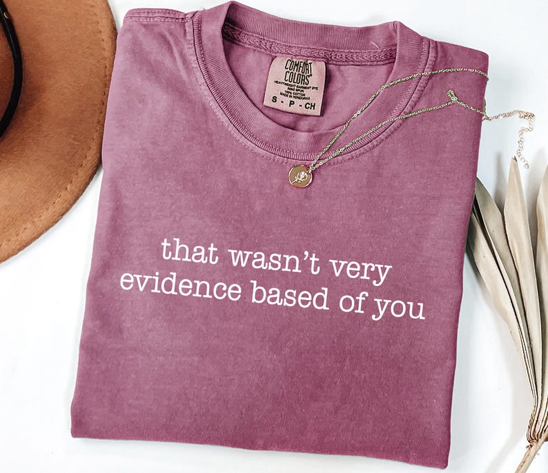 May include: A mauve Comfort Colors t-shirt with the phrase "that wasn't very evidence based of you" in white text. A gold necklace with a pendant and a brown hat are also visible.