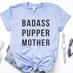 May include: A light blue t-shirt with the text "BADASS PUPPER MOTHER" printed in black.