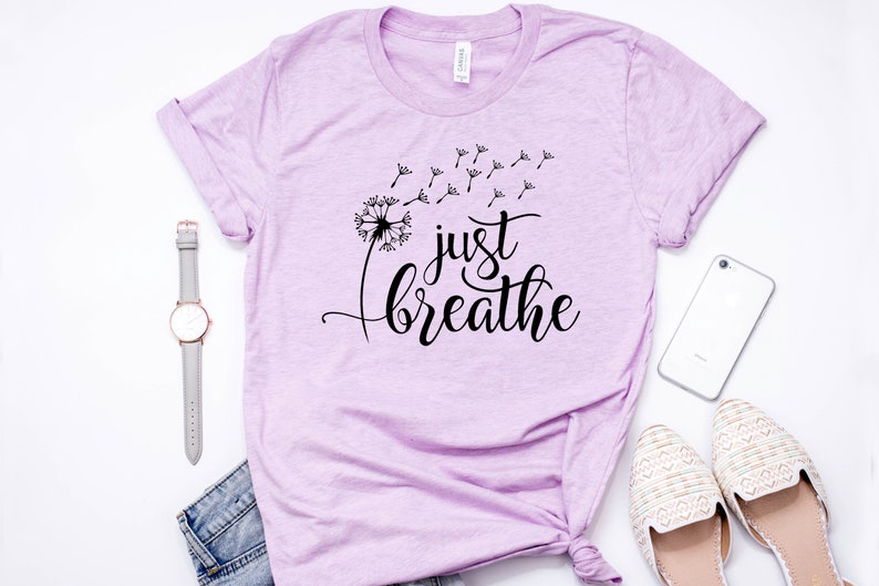 Op de afbeelding: Een lichtpaars T-shirt met een zwarte paardenbloem afbeelding en de tekst "just breathe" in een cursief schrift.