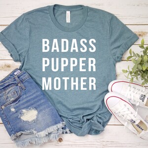 May include: A teal blue t-shirt with the text "BADASS PUPPER MOTHER" in white lettering.