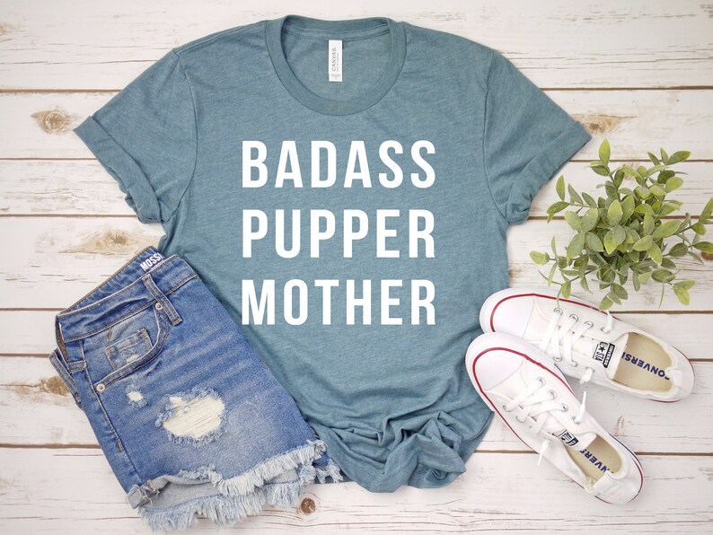 May include: A teal blue t-shirt with the text "BADASS PUPPER MOTHER" in white lettering.