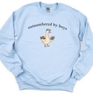 May include: Light blue sweatshirt with the text "outnumbered by boys" above a cartoon goose wearing a hat and holding a football and a toy truck. The sweatshirt has long sleeves and a crew neck.