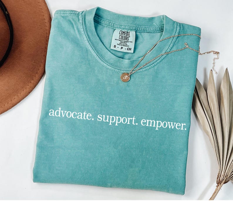 Op de afbeelding: Een turquoise Comfort Colors t-shirt met de woorden "advocate. support. empower." in het wit. Een gouden ketting met een hanger ligt op het shirt. Een bruine hoed en gedroogde bladeren zijn op de achtergrond.