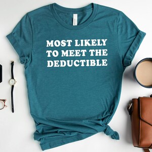 May include: Teal t-shirt with the white text "MOST LIKELY TO MEET THE DEDUCTIBLE." The shirt is styled with accessories including a watch, glasses, earrings, a coffee mug, a purse, and a small plant.