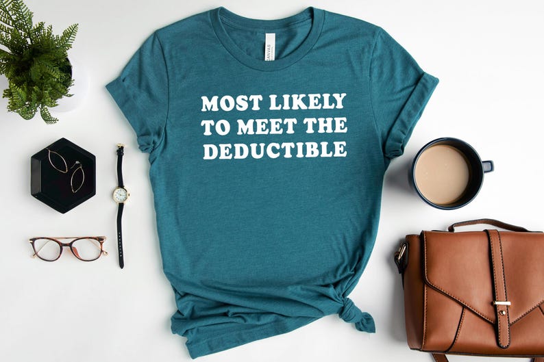 May include: Teal t-shirt with the white text "MOST LIKELY TO MEET THE DEDUCTIBLE." The shirt is styled with accessories including a watch, glasses, earrings, a coffee mug, a purse, and a small plant.