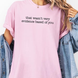 May include: A light pink t-shirt with the phrase "that wasn't very evidence based of you" printed in black. The shirt is being worn with a denim jacket.