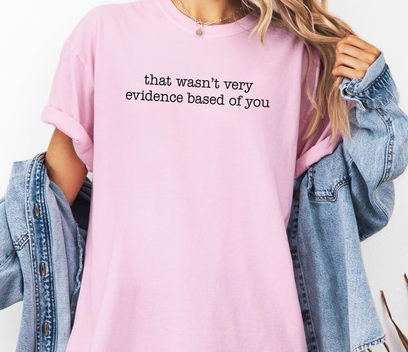 May include: A light pink t-shirt with the phrase "that wasn't very evidence based of you" printed in black. The shirt is being worn with a denim jacket.