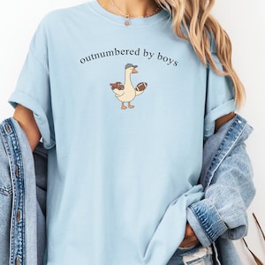 May include: Pale blue t-shirt featuring the phrase "outnumbered by boys" and a cartoon goose with a hat, football, and toy truck. The shirt has a crew neck and short sleeves.