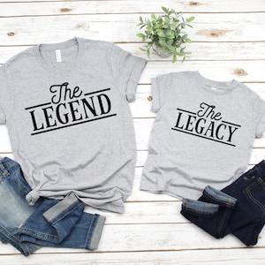 May include: Two gray t-shirts with black text. The larger shirt says "The Legend" and the smaller shirt says "The Legacy".