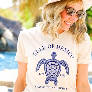 Puede incluir: Camiseta color crema con un gr&aacute;fico azul de una tortuga marina y el texto "GULF OF MEXICO" encima. Debajo de la tortuga est&aacute; "EST. 1550" y "STAY SALTY AND RESIST". La camiseta tiene mangas cortas.