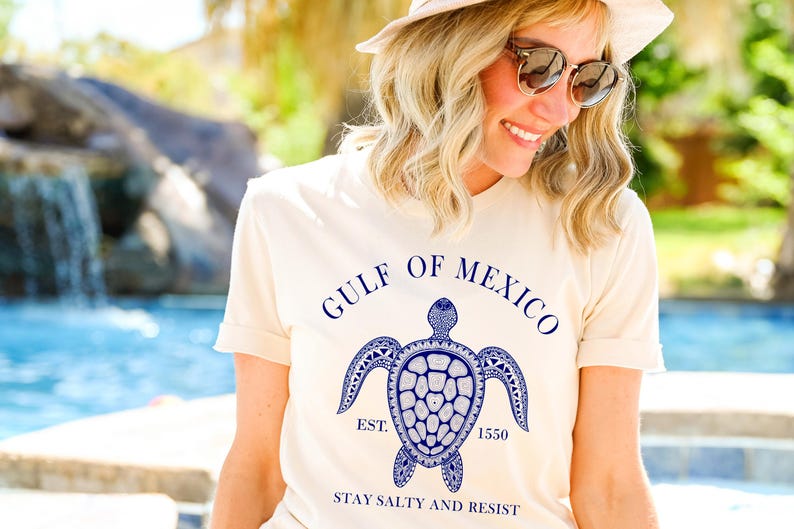Puede incluir: Camiseta color crema con un gr&aacute;fico azul de una tortuga marina y el texto "GULF OF MEXICO" encima. Debajo de la tortuga est&aacute; "EST. 1550" y "STAY SALTY AND RESIST". La camiseta tiene mangas cortas.