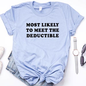 May include: A light blue t-shirt with the text "MOST LIKELY TO MEET THE DEDUCTIBLE" in black. The shirt is styled with a pair of white flats, denim shorts, a watch, and sunglasses.