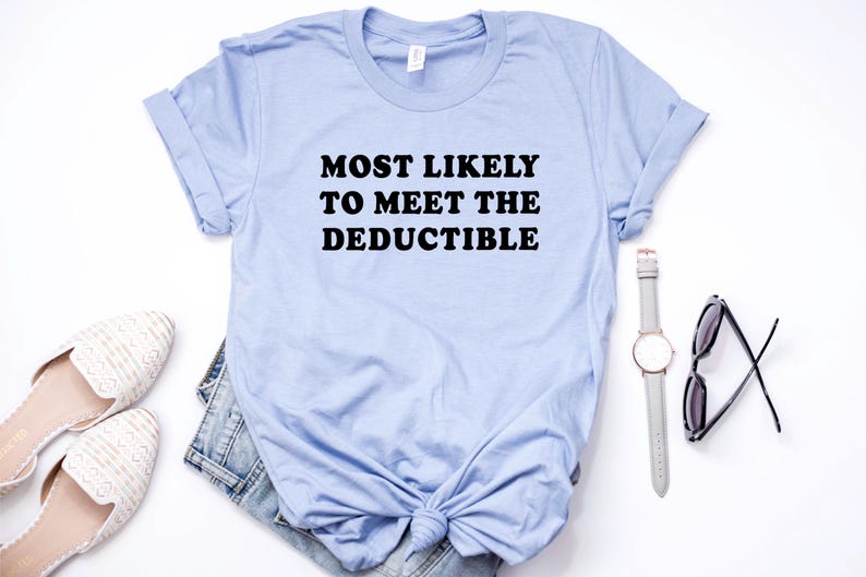 May include: A light blue t-shirt with the text "MOST LIKELY TO MEET THE DEDUCTIBLE" in black. The shirt is styled with a pair of white flats, denim shorts, a watch, and sunglasses.
