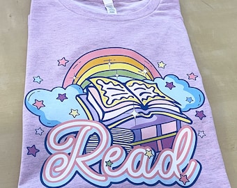 Puede incluir: Una camiseta morada con un arcoíris, una pila de libros y la palabra "Leer" en una fuente caprichosa. El diseño incluye estrellas y nubes.