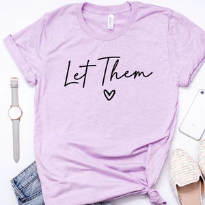 May include: A lavender t-shirt with the text "Let Them" in black script with a small heart underneath.