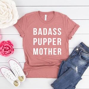 May include: A pink t-shirt with the text "BADASS PUPPER MOTHER" in white lettering. The shirt is laid flat on a white wooden surface with a pair of white sneakers, blue jeans, and pink and white flowers.