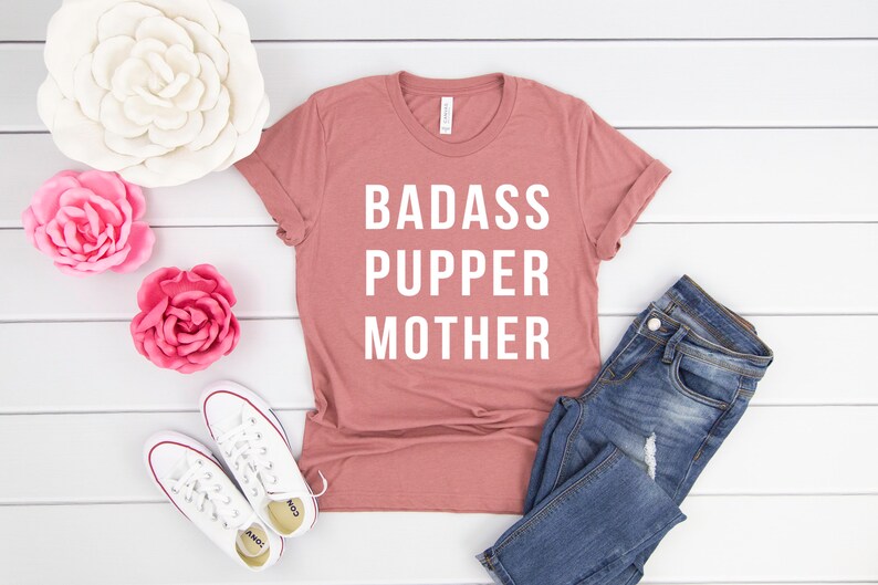 May include: A pink t-shirt with the text "BADASS PUPPER MOTHER" in white lettering. The shirt is laid flat on a white wooden surface with a pair of white sneakers, blue jeans, and pink and white flowers.