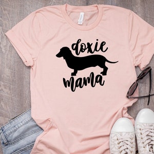 May include: A pink t-shirt with a black silhouette of a dachshund and the text "doxie mama".