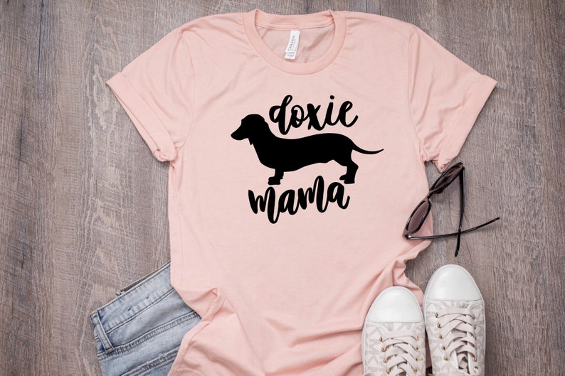 May include: A pink t-shirt with a black silhouette of a dachshund and the text "doxie mama".