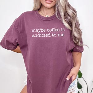 May include: A maroon t-shirt with the phrase "maybe coffee is addicted to me" printed in white. The shirt has short sleeves and a relaxed fit, suitable for casual wear.