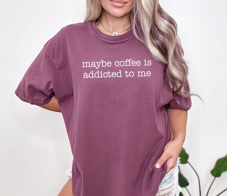 May include: A maroon t-shirt with the phrase "maybe coffee is addicted to me" printed in white. The shirt has short sleeves and a relaxed fit, suitable for casual wear.