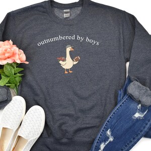 May include: Dark gray sweatshirt with the text "outnumbered by boys" above a cartoon goose wearing a hat and holding a football. The sweatshirt is paired with blue jeans and white shoes.